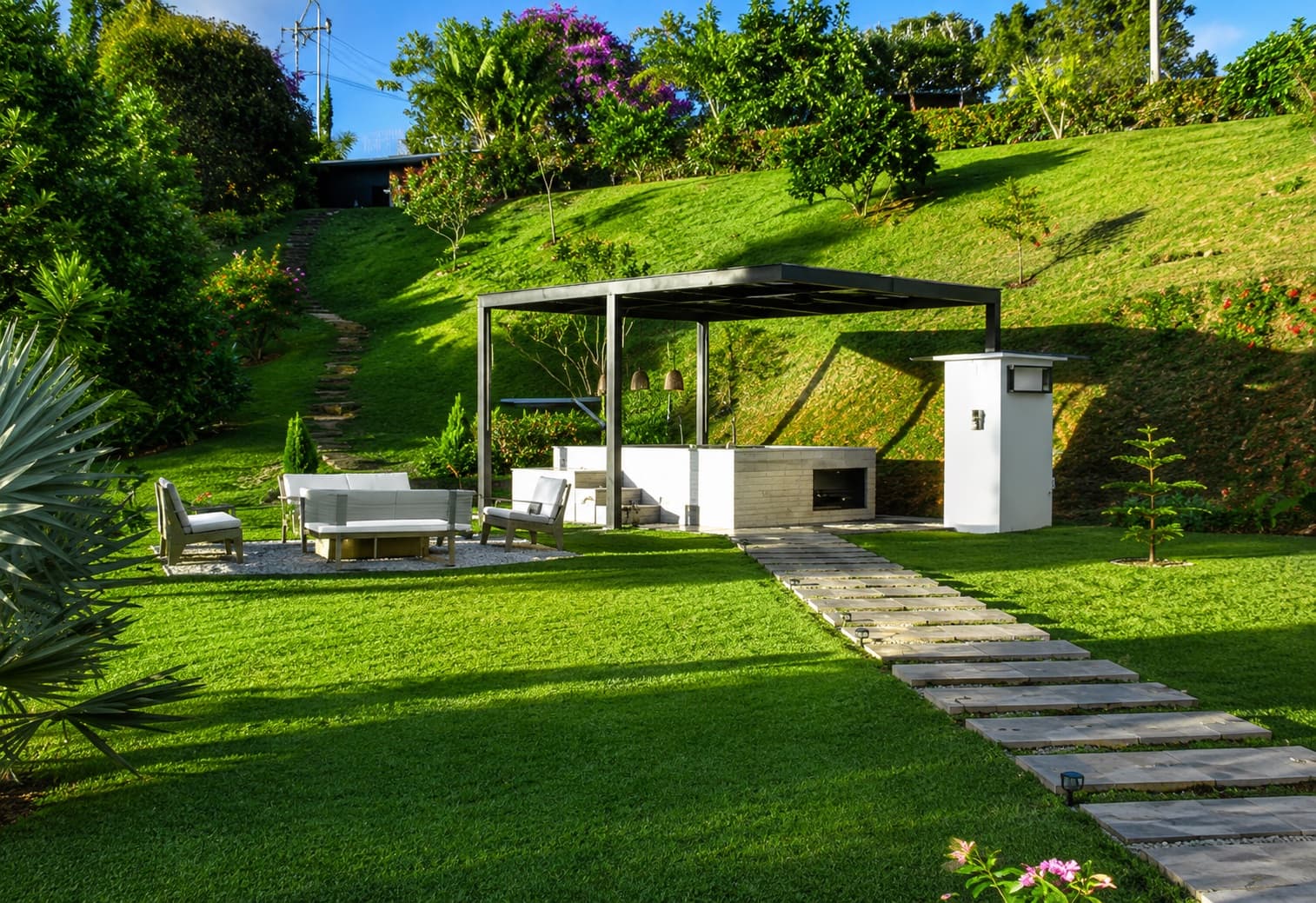 Sala exterior con pérgola y zona BBQ entre jardines